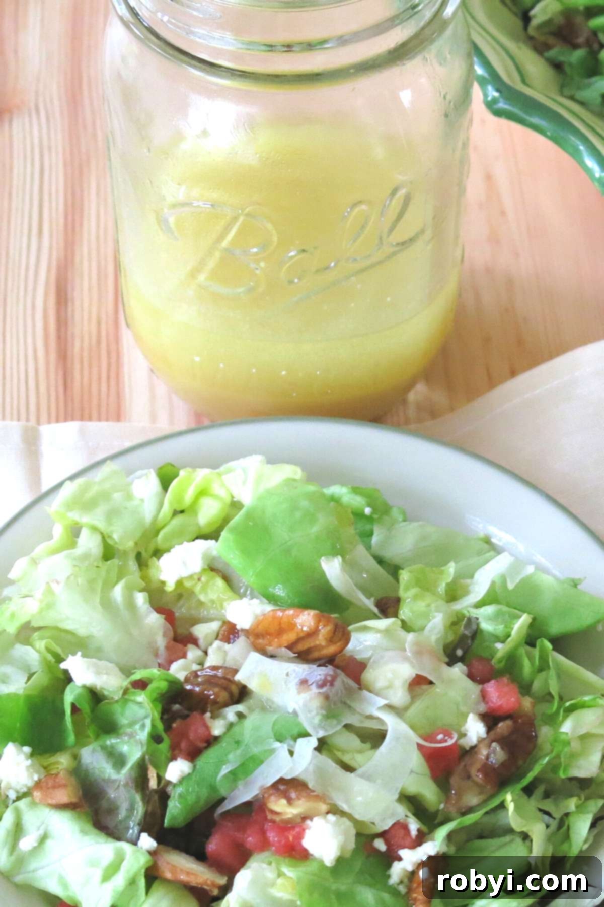 A mason jar filled with bright Orange Honey Mustard Vinaigrette, positioned behind a serving plate of the complete Summer Fennel Salad, showcasing the shaved fennel, watermelon, feta, and candied pecans. The scene highlights the dressing as a key component of the dish.