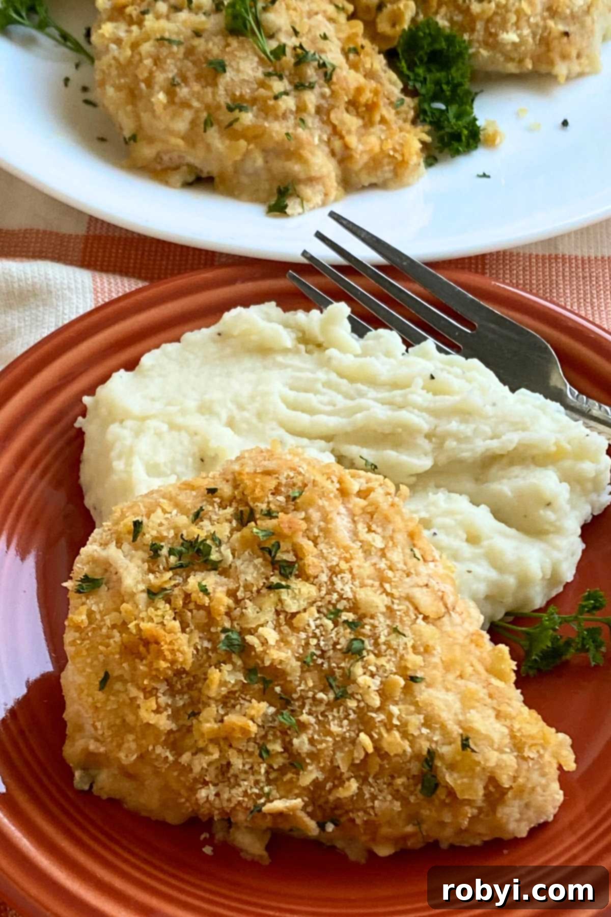 Crispy Ritz Cracker Chicken Perfection 9 Serving of baked chicken coated with Ritz cracker crumb topping on top of creamy cauliflower mash on plate with a fork with a platter with more chicken behind it.