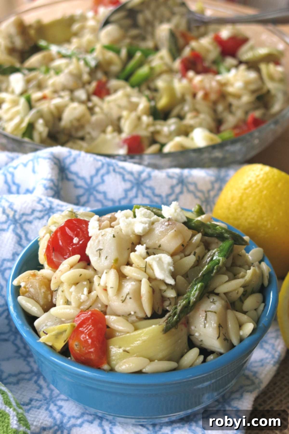 Pan-Seared Scallops over Warm Orzo with Marinated Artichokes and Feta 4 Mediterranean warm orzo salad with scallops, asparagus, artichokes, tomatoes, and feta cheese in a bowl, with serving dish behind it.