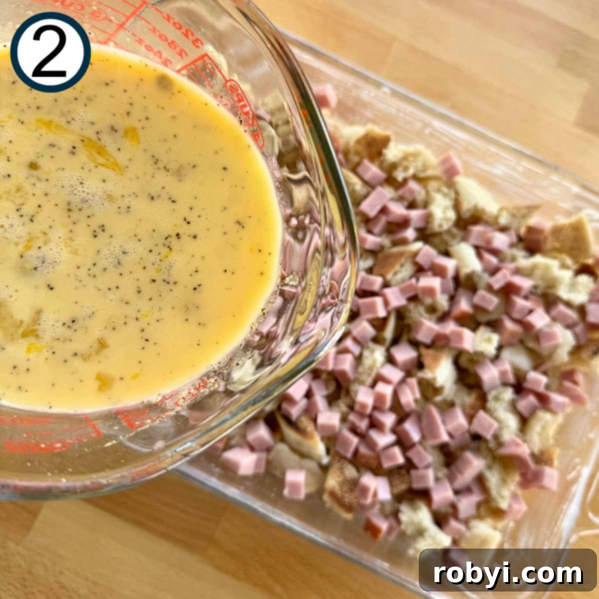 Egg mixture being poured over pieces of English muffins layered with diced ham in a large glass baking dish.