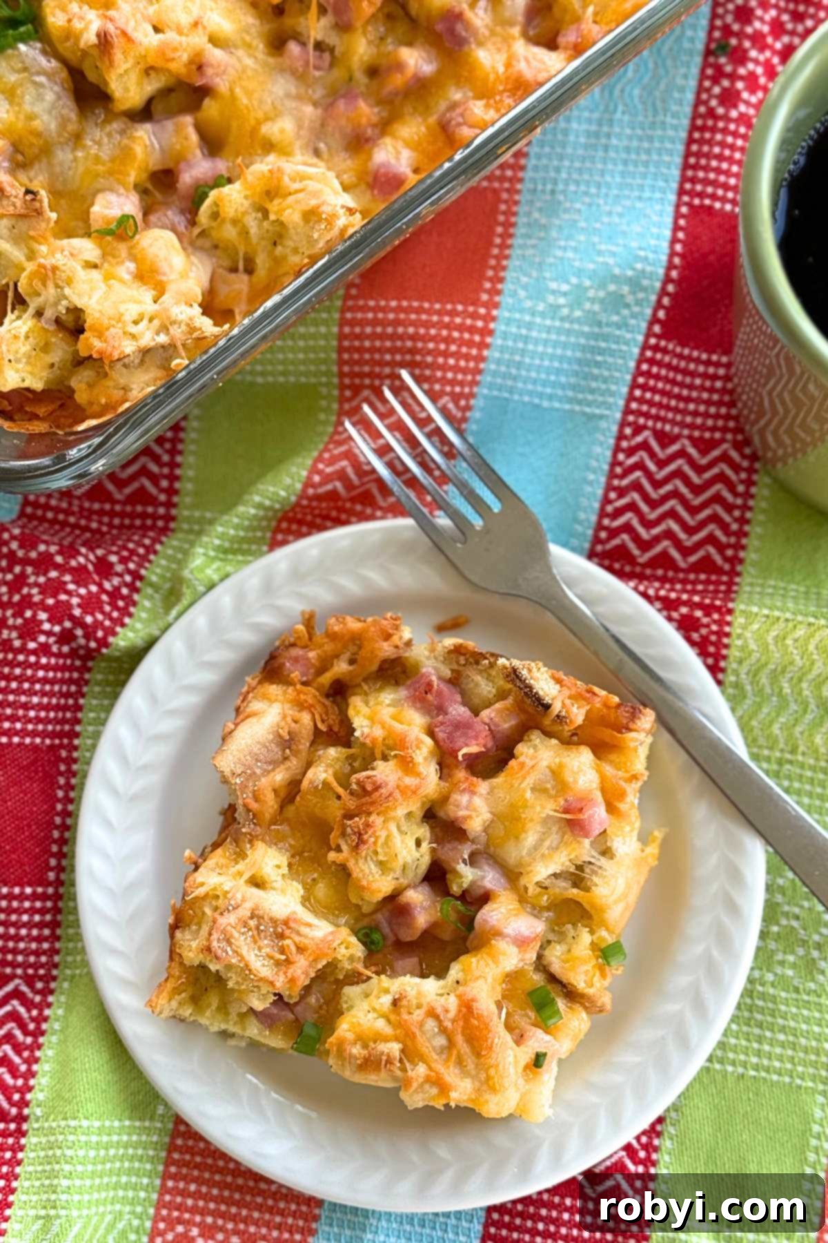 Serving of ham and cheese strata on a white plate with a fork with the casserole dish and green coffee mug behind it.