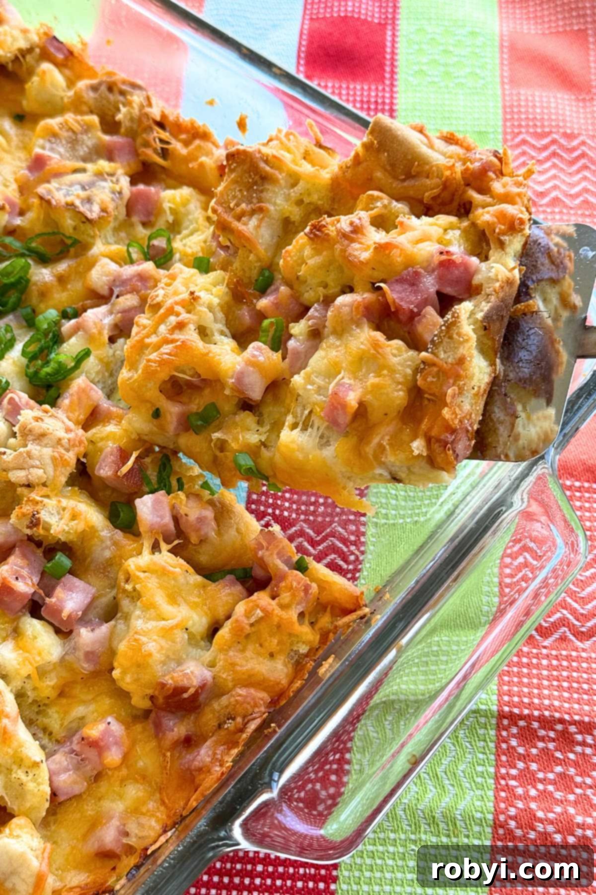 Spatula lifting a serving of ham and cheese strata our of a glass baking dish that's on a pastel colored cloth napkin.