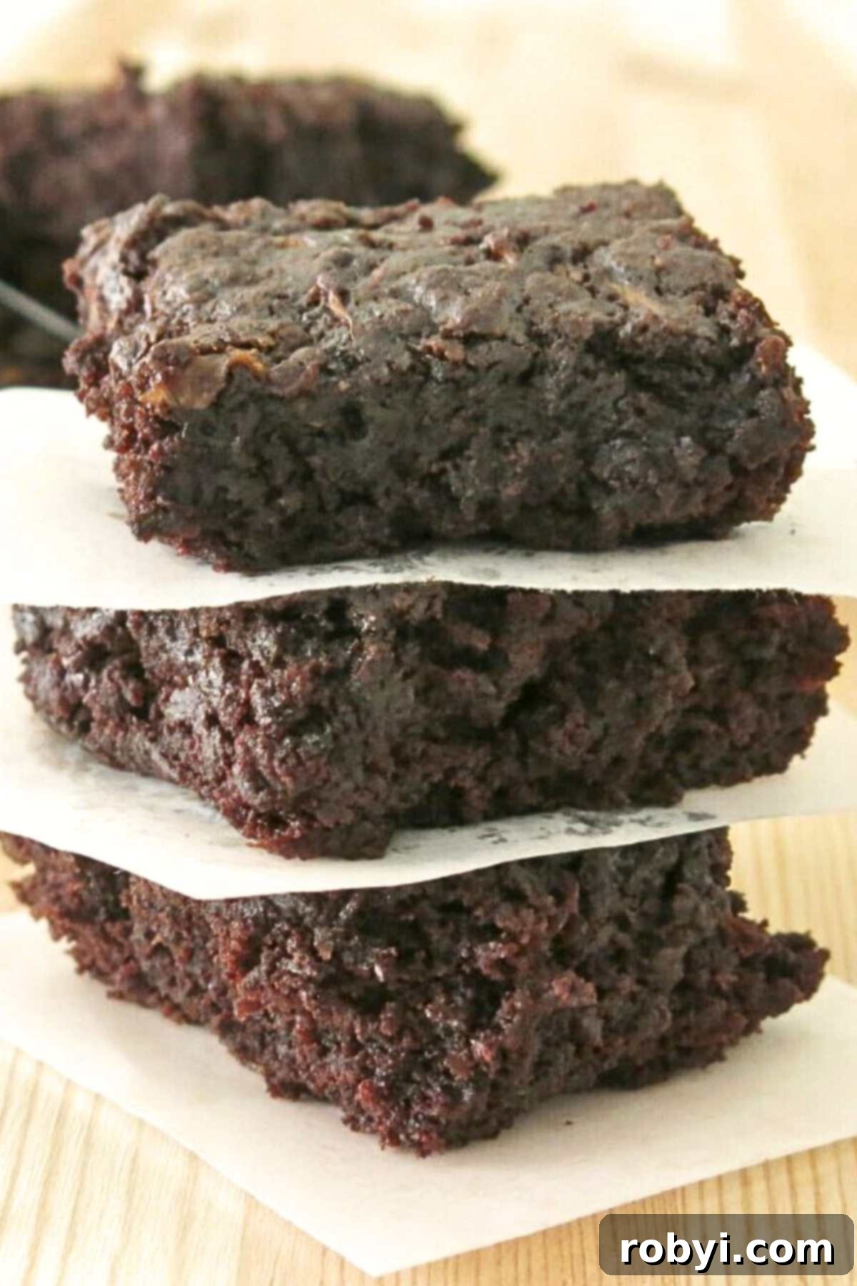 Decadent Zucchini Chocolate Brownies 2 A tantalizing stack of three fudgy zucchini brownies, perfectly separated by parchment paper, resting on a rustic butcher block table top. Each brownie boasts a rich, dark chocolate hue and a delightfully dense, moist texture.