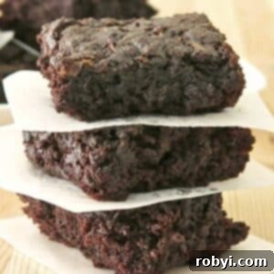Decadent Zucchini Chocolate Brownies 11 A stack of three irresistibly fudgy zucchini brownies, neatly separated by parchment paper, resting on a textured butcher block surface.