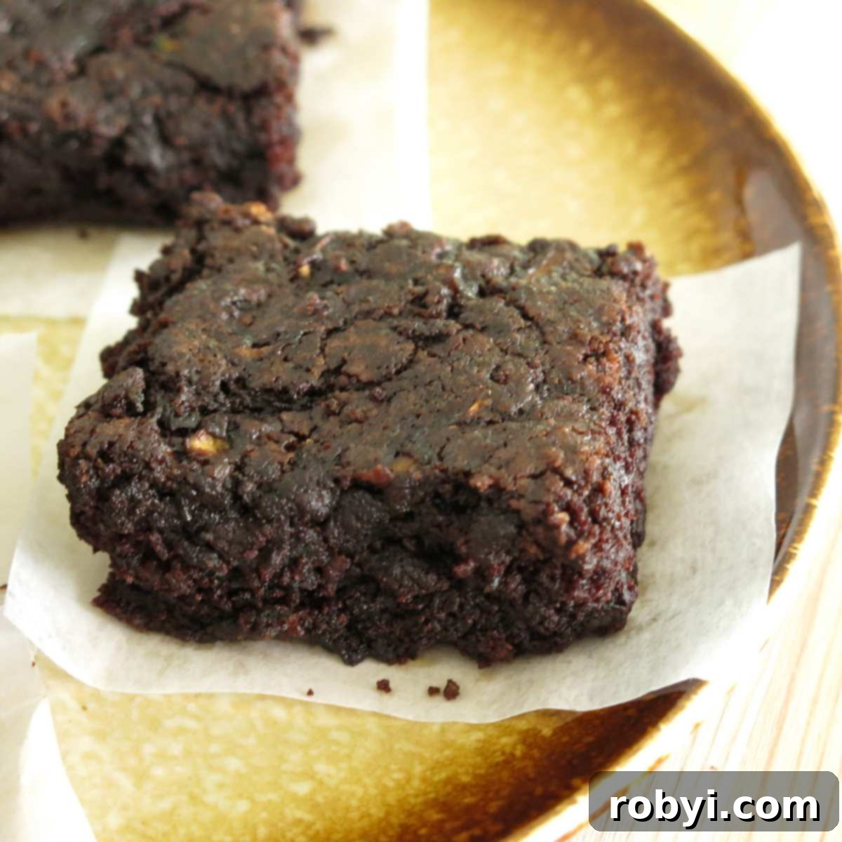 Decadent Zucchini Chocolate Brownies 10 A single delectable fudgy zucchini brownie rests invitingly on a white plate, with another partially visible behind it, showcasing its rich chocolate color and moist interior.