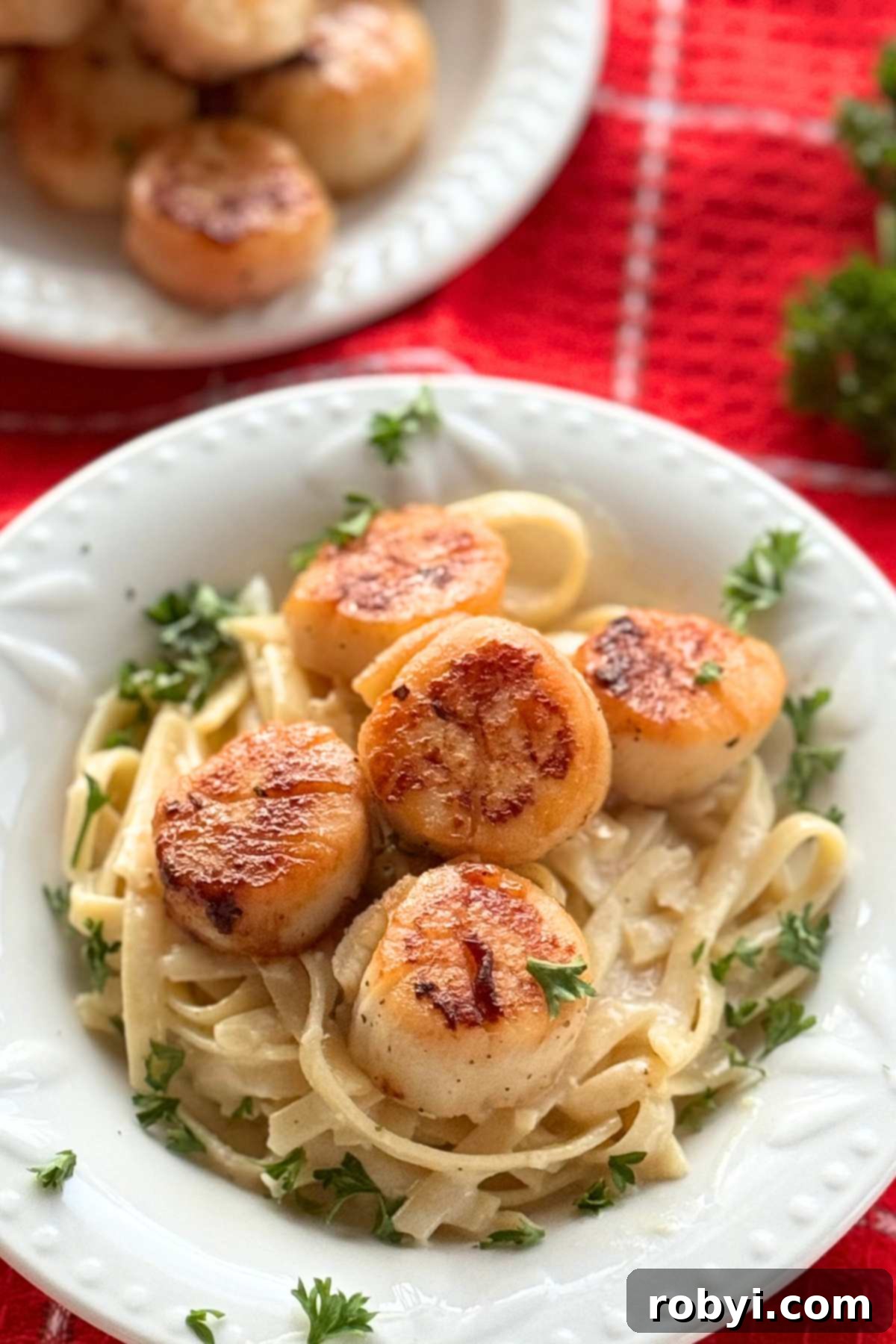 Quick and Creamy Scallop Fettuccine Alfredo 7 Bowl with creamy fettuccine alfredo topped with seared-scallops and parsley with a plate with more cooked scallops behind it.