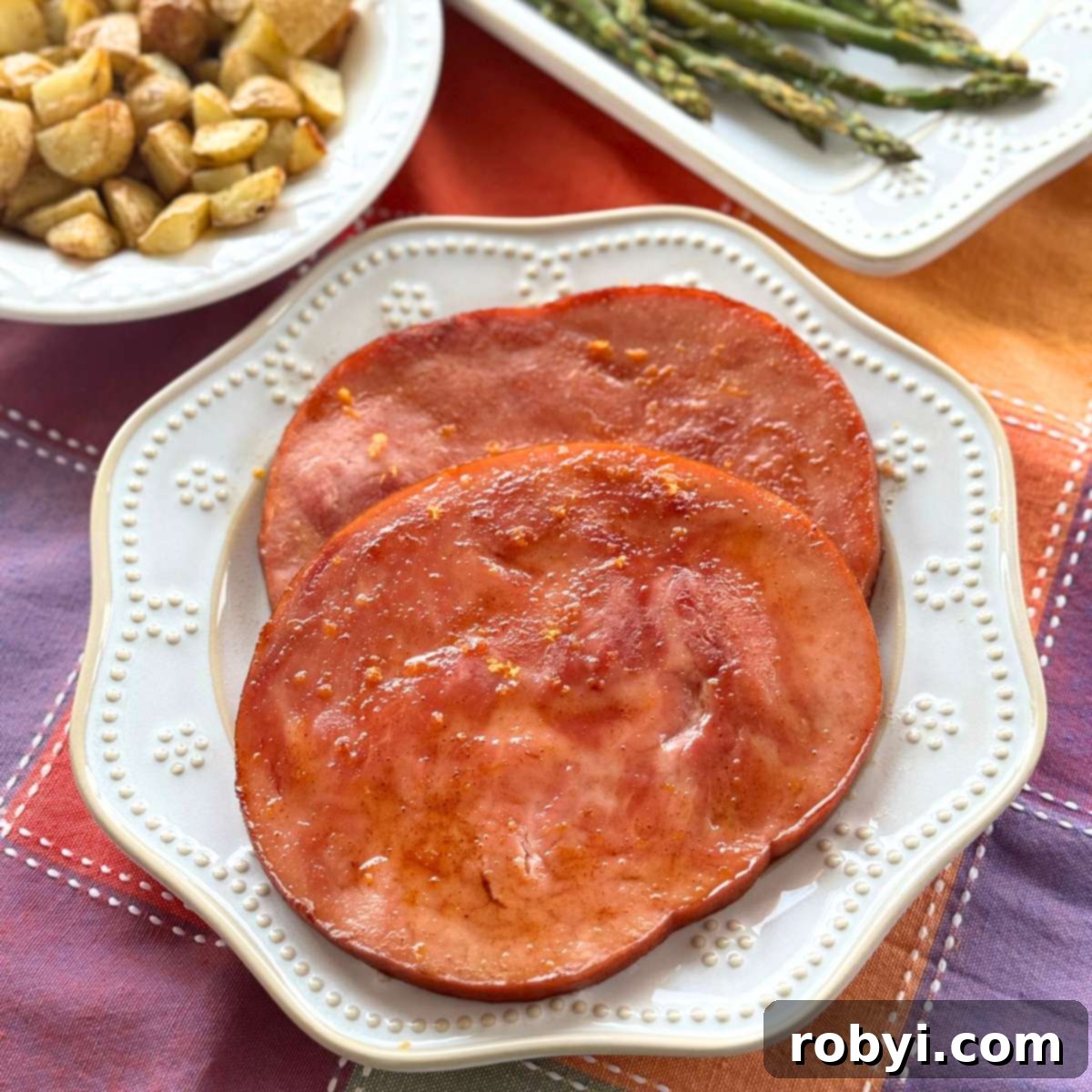 Two baked ham steaks coated with a honey, orange juice, brown sugar glaze on a white plate with asparagus and roasted potatoes behind it.