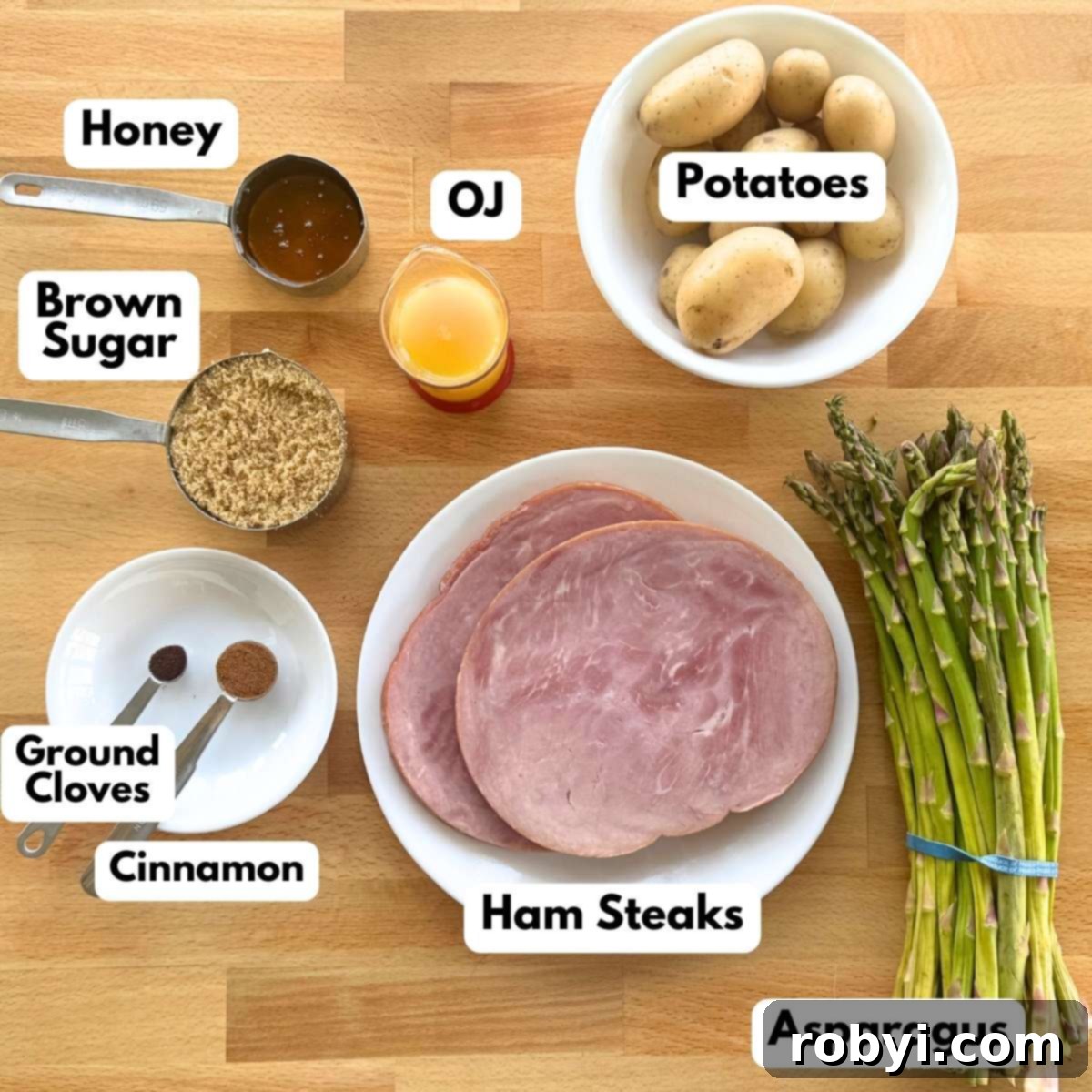 Two ham steaks, honey, orange juice, brown sugar, cinnamon, ground cloves, asparagus, and small potatoes on a cutting board.