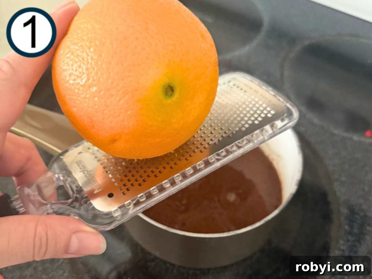 Orange being zested over a small sauce pot with a brown mixture of brown sugar, orange juice, honey, and spices.