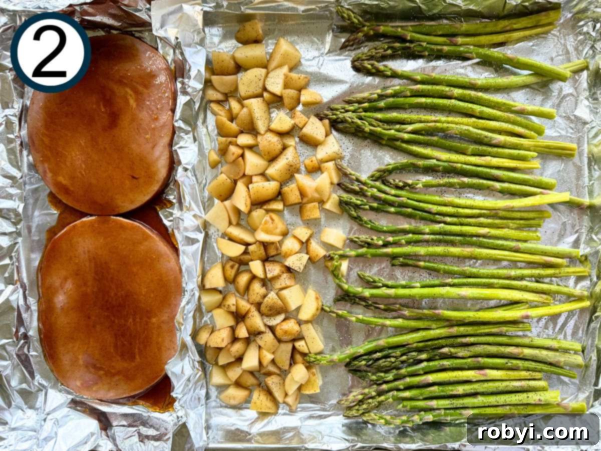 Uncooked ham steaks brushed with a honey orange glaze, diced pototes, and asparagus spears on a large sheet pan lined with aluminum foil.