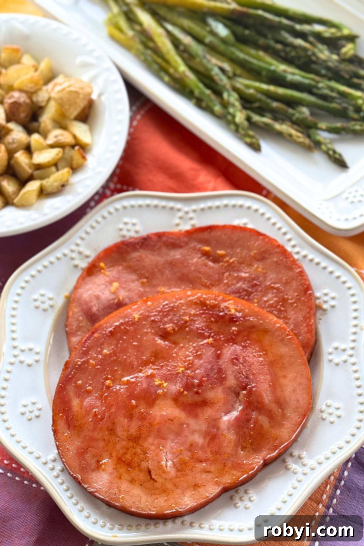 Two glazed ham steaks on a plate with asparagus and roasted potatoes in dishes behind it.