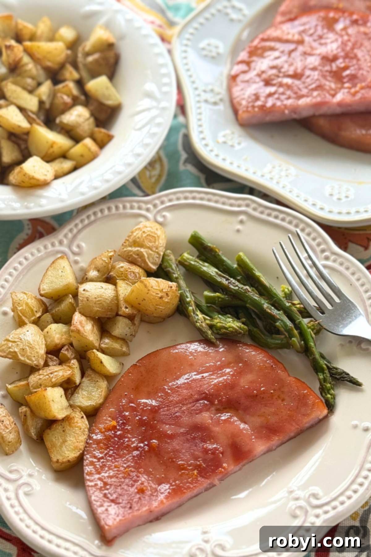 Serving of ham steak, roasted potatoes, and asparagus on a plate with more potatoes and slices of ham in the background.