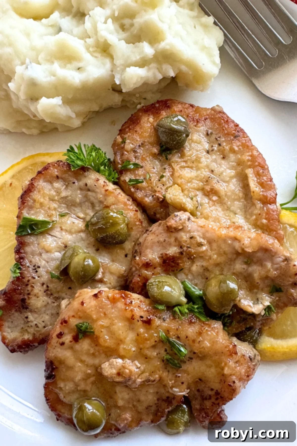 Four pork tenderloin piccata medallions topped with lemon butter sauce, capers, and fresh parsley on a plate with mashed potatoes and a fork in the background.