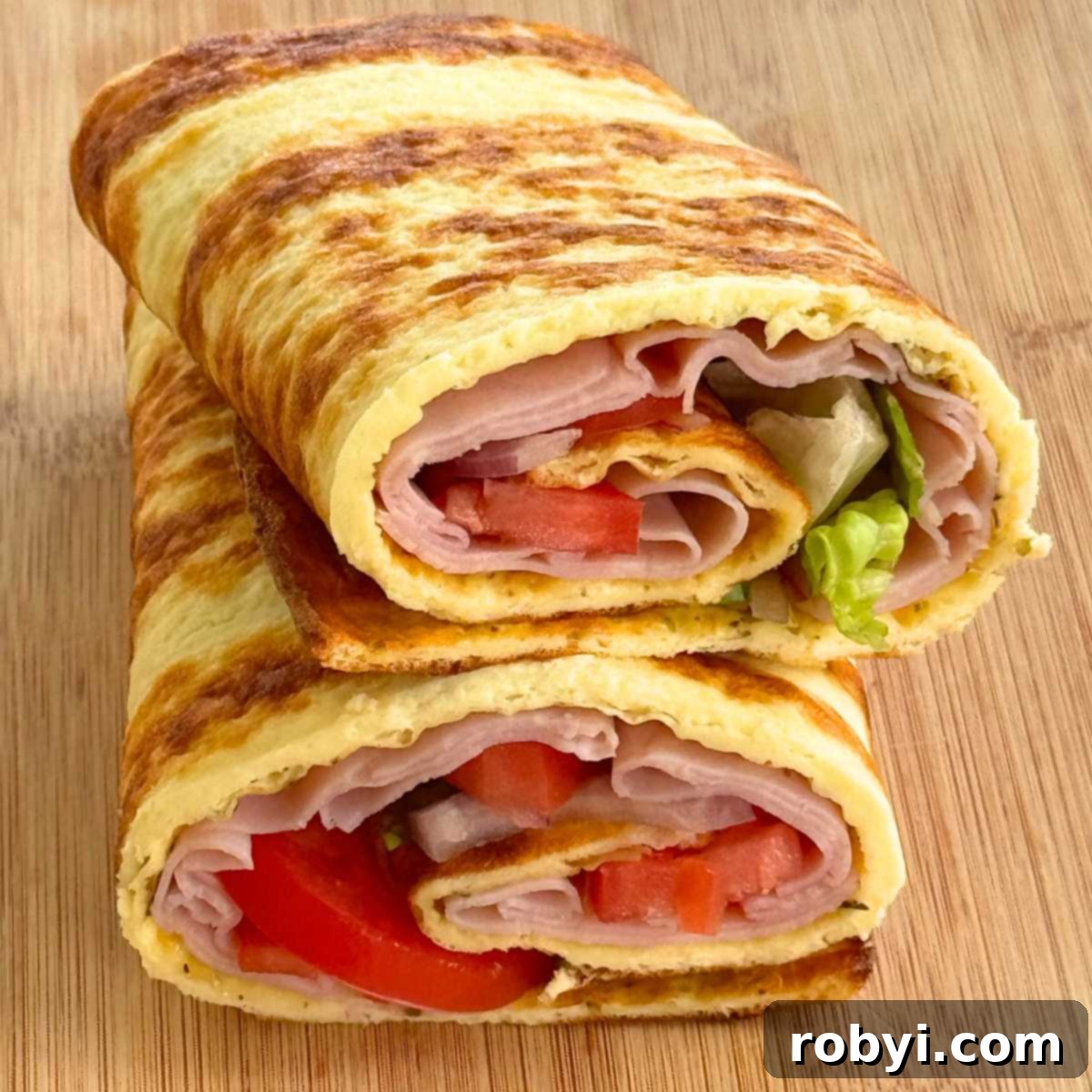 Cottage cheese flatbread, with ham, tomatoes, onion, and lettuce rolled inside, cut in half and stacked on a cutting board.