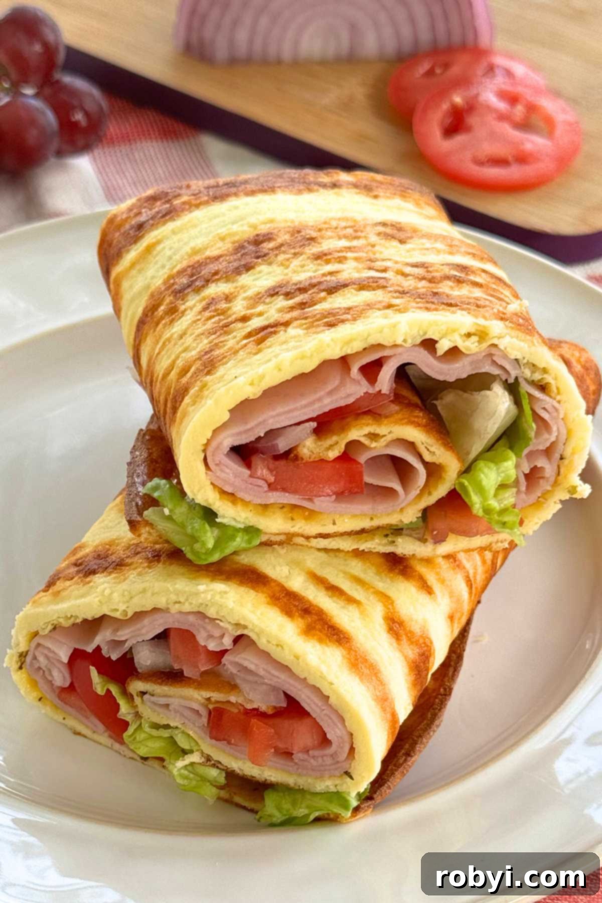 Cottage cheese flatbread, with ham, tomatoes, onion, and lettuce rolled inside, cut in half and stacked on a plate with a cutting board behind it with tomato slices and onion.