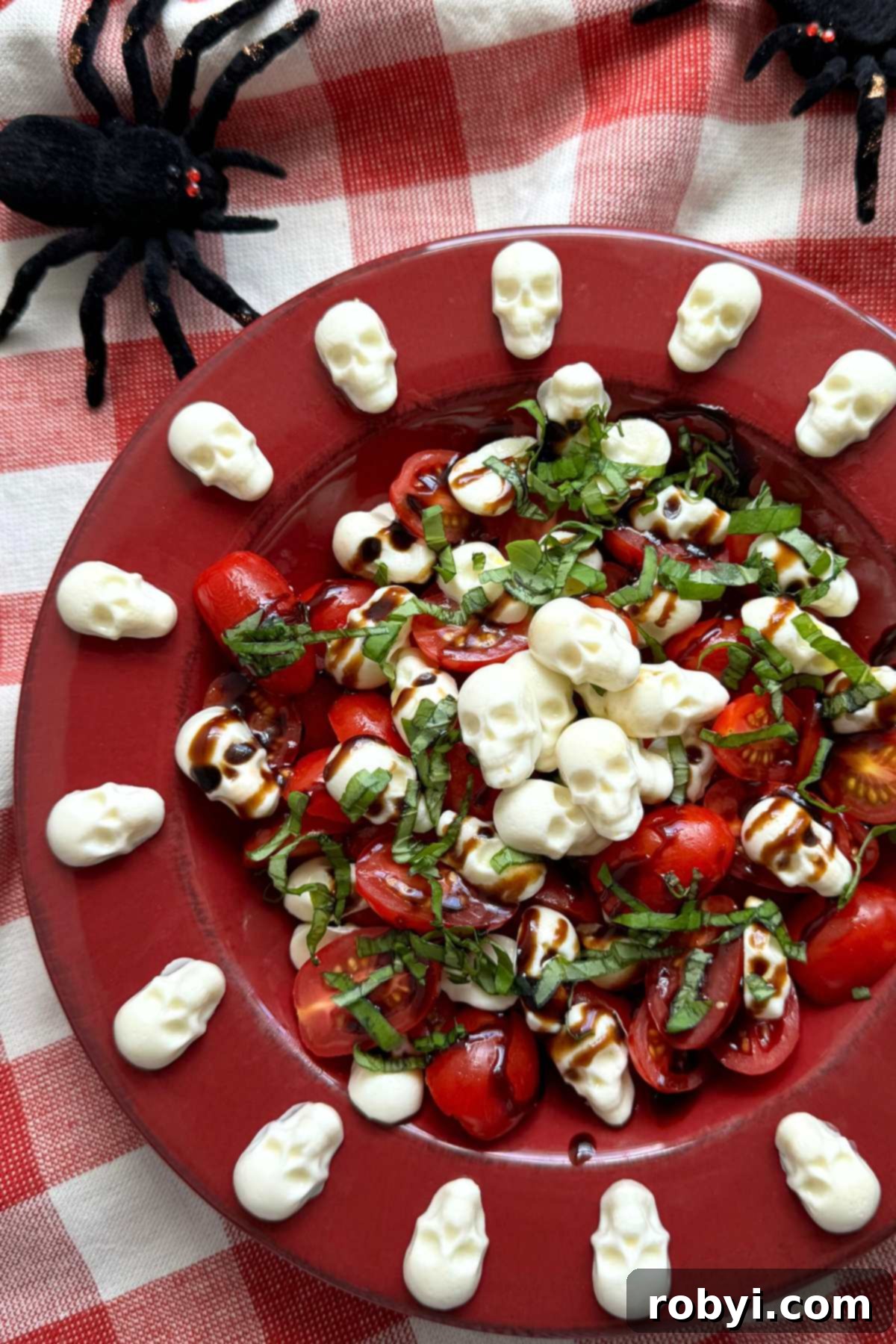 A large, vibrant red bowl filled with the Halloween Caprese Salad, adorned with mozzarella skulls arranged around the flat edge and piled in the center, with decorative spiders in the background adding to the spooky ambiance.