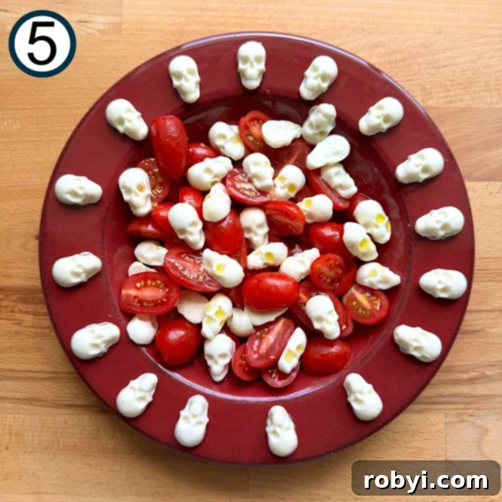 A red plate beautifully arranged with halved grape tomatoes and delicate mozzarella skulls, forming the foundation of the Caprese salad.