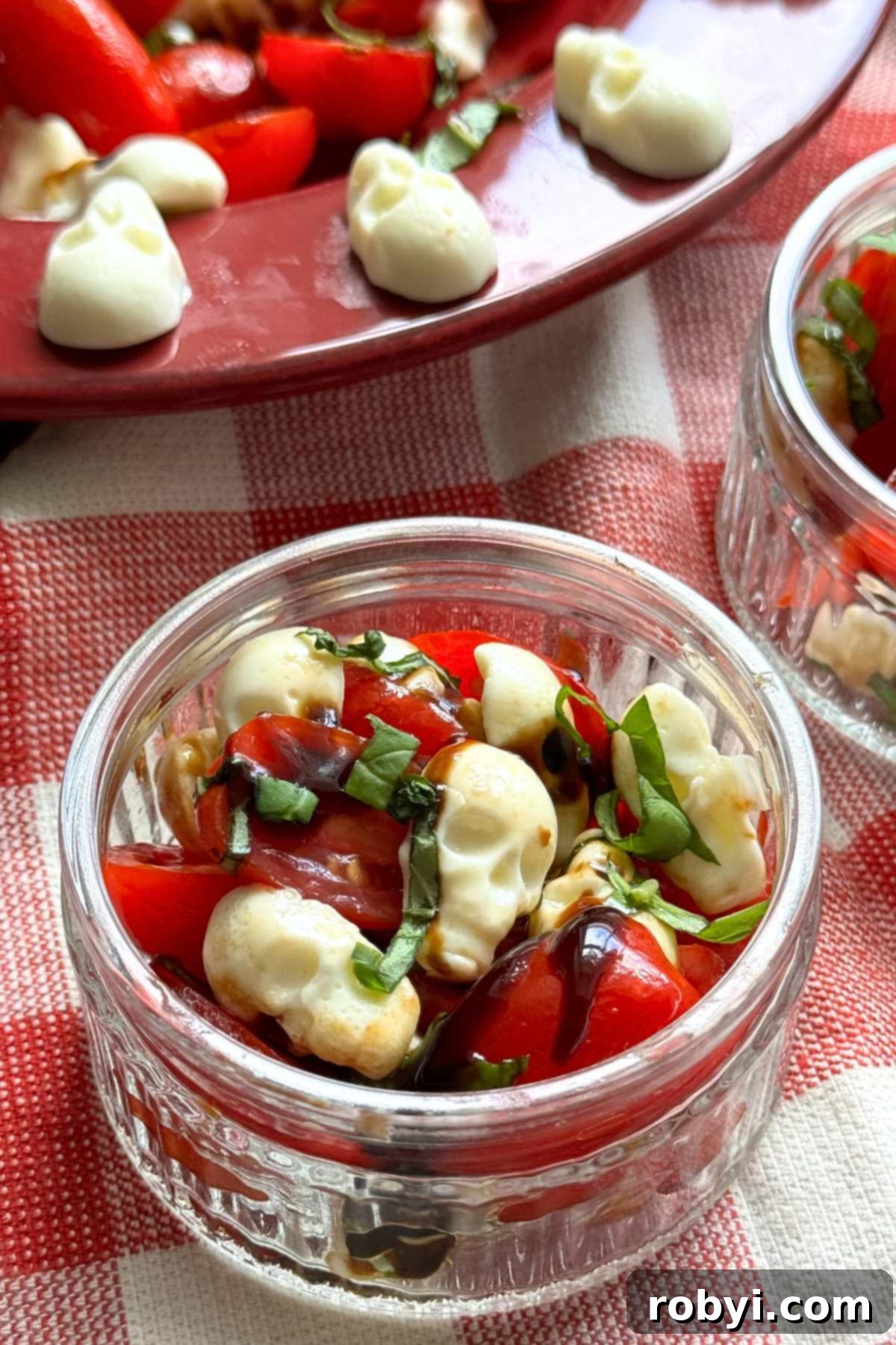 A small glass bowl holding a single serving of Halloween Caprese Salad with spooky mozzarella skulls, positioned in front of a larger bowl showcasing the full salad, creating a delightful visual depth.