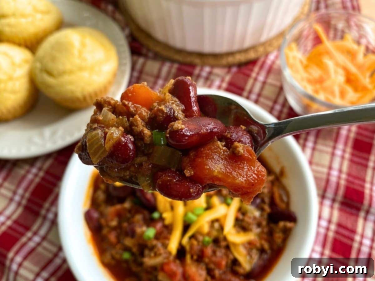 A spoon expertly lifting a generous portion of easy Southern chili from a bowl, with warm corn muffins and grated cheese artfully blurred in the background.