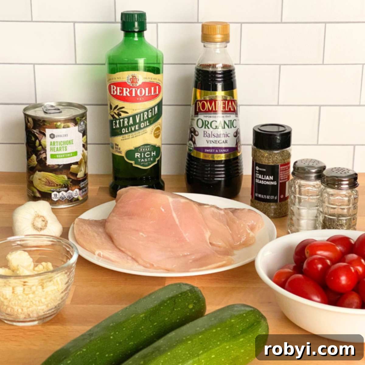 Tek Kapta Pratik Akdeniz Fırın Tavuğu 3 Ingredients laid out for Mediterranean Chicken Bake: boneless, skinless chicken breasts, zucchini, grape tomatoes, a can of artichoke hearts, olive oil, balsamic vinegar, Italian seasoning, fresh garlic, salt, pepper, and crumbled feta cheese.
