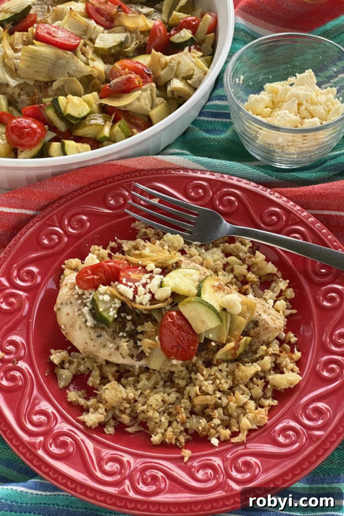 Tek Kapta Pratik Akdeniz Fırın Tavuğu 5 Mediterranean baked chicken topped with crumbled feta cheese on a vibrant red plate, with a larger platter of the dish and a cup of feta cheese in the background.