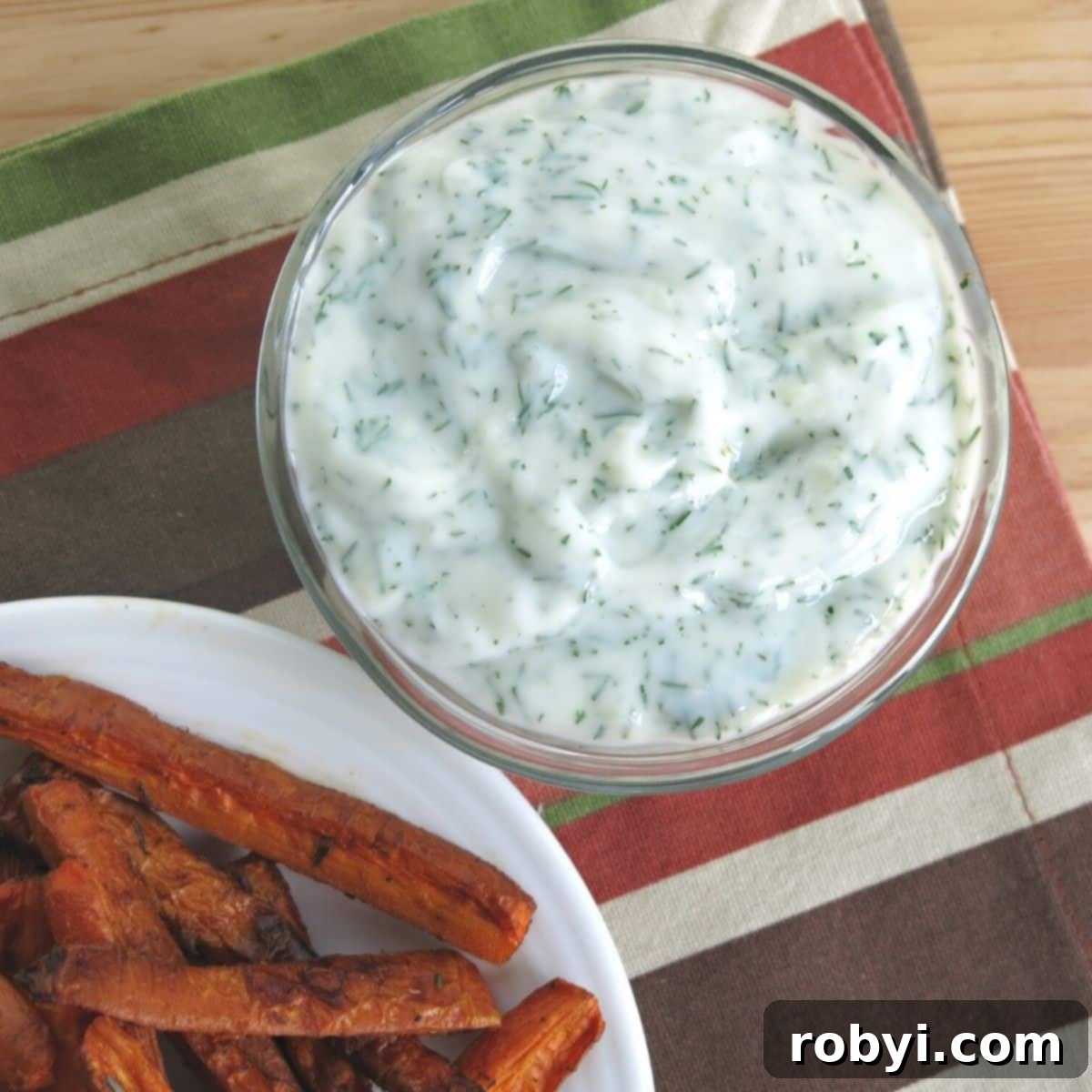 A bowl of creamy yogurt dill dipping sauce placed next to a plate of healthy, crispy carrot fries.