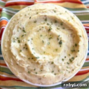Creamy crock-pot mashed potatoes with cream cheese and sour cream in a serving bowl with pools of butter, and chives on top.