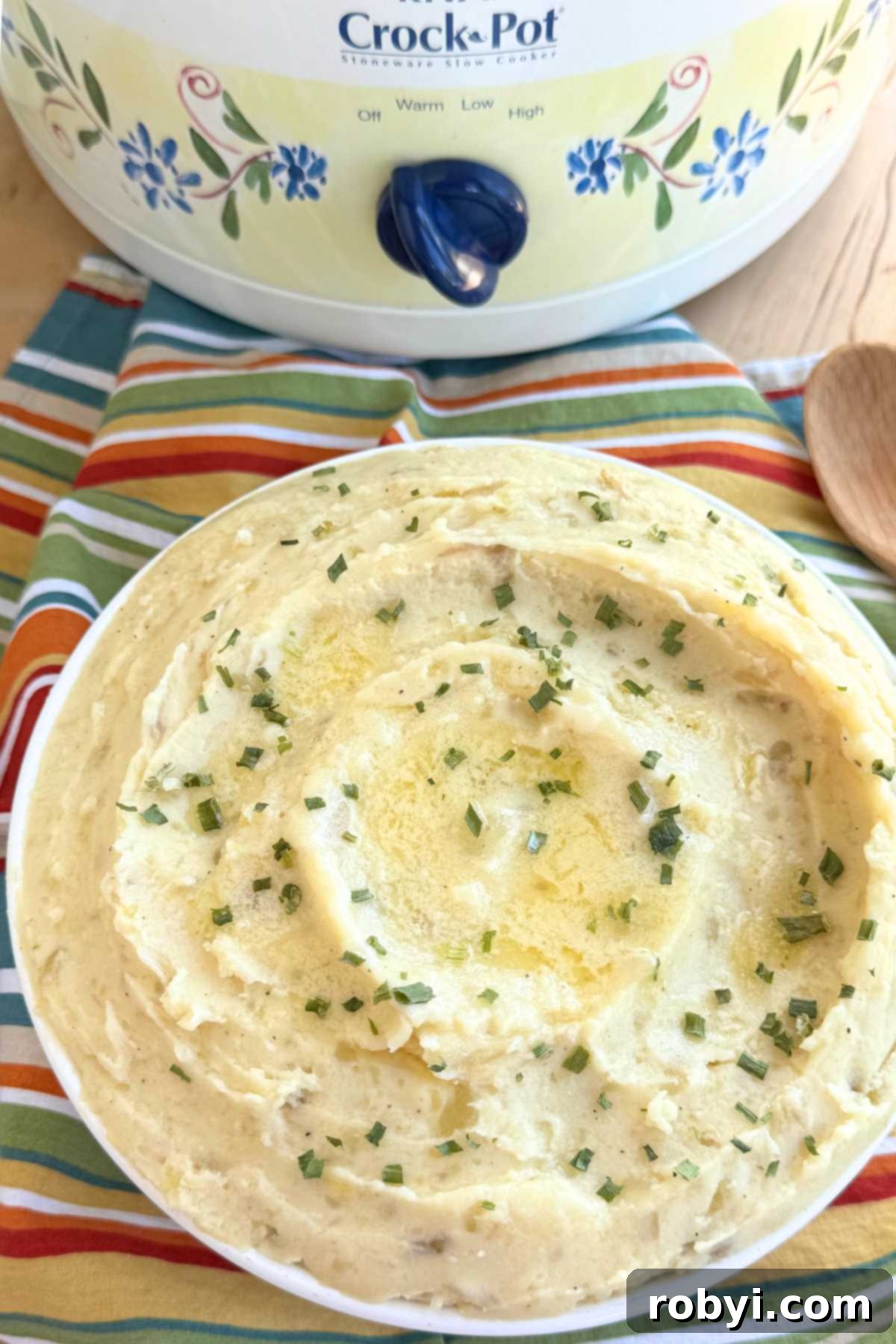 Creamy slow cooker mashed potatoes topped with melted butter and chives in a large bowl with a slow cooker behind it.