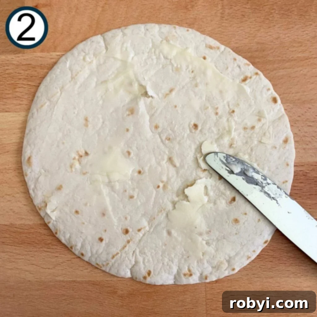 A hand expertly spreading a thin layer of butter onto a flour tortilla, preparing it for pan-frying.