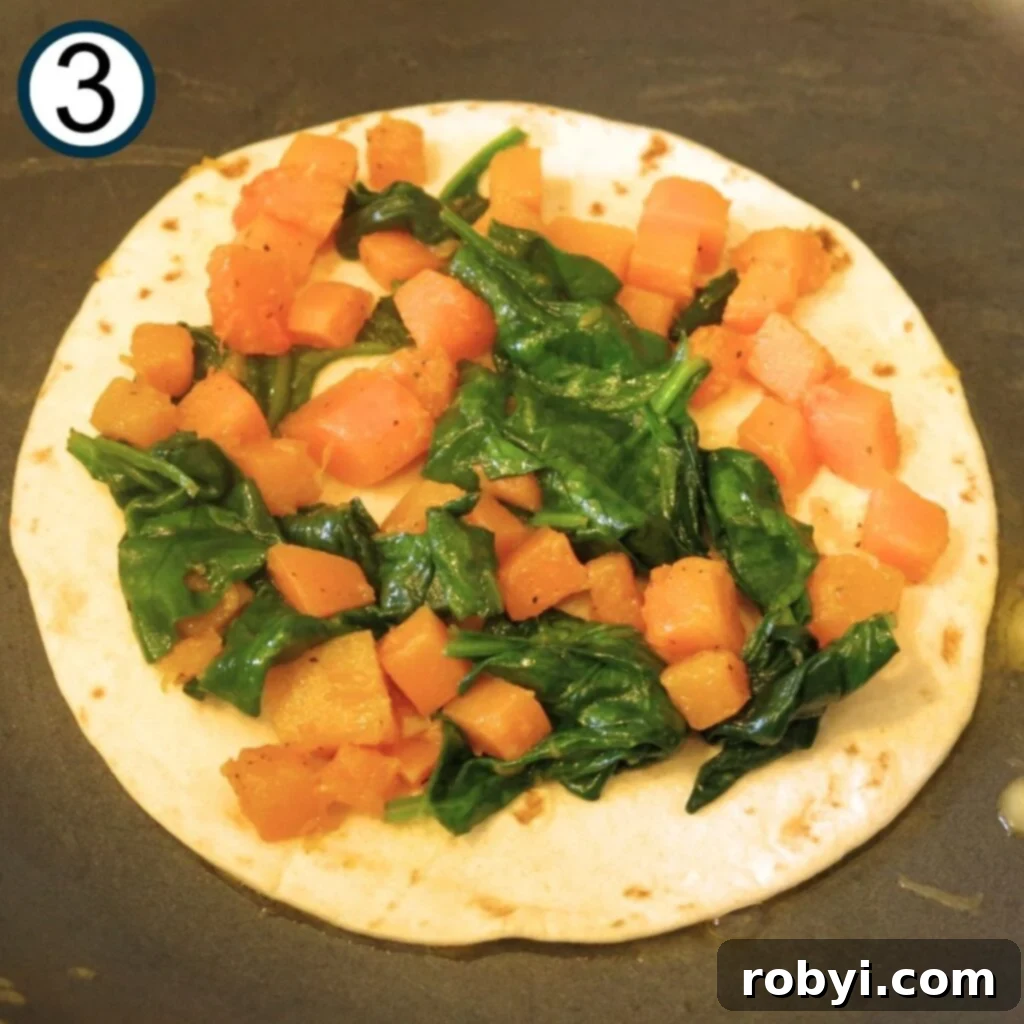 A half-assembled butternut squash quesadilla in a hot skillet, with a tortilla base, a layer of squash and spinach, and shredded cheese awaiting its top layer.