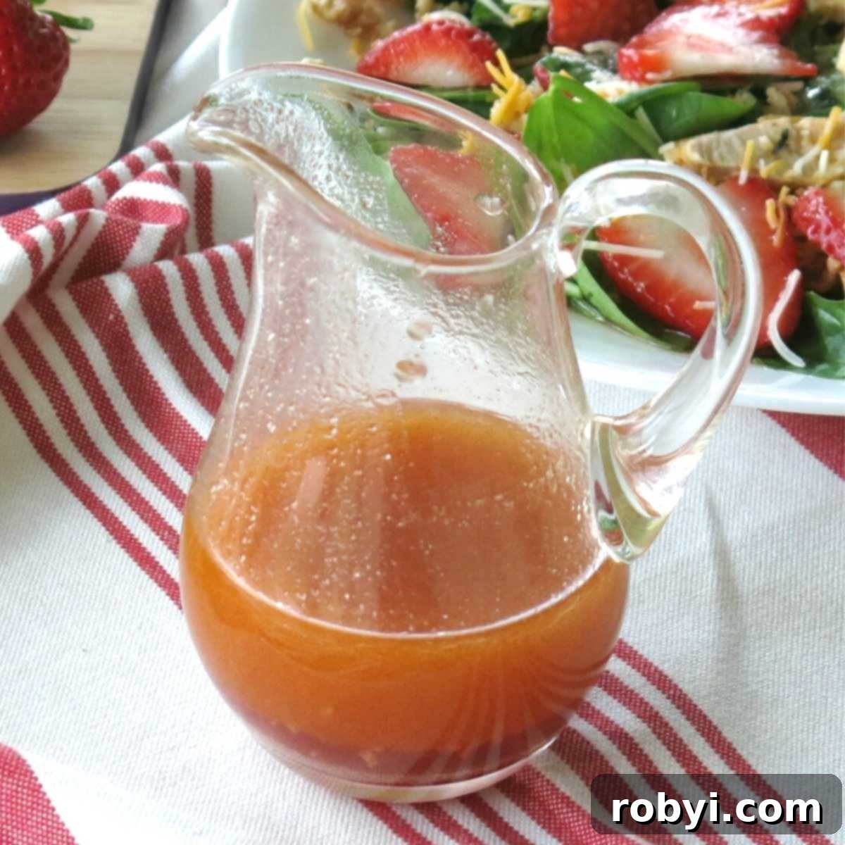A glass container filled with vibrant Sweet Red Wine Vinaigrette Dressing, ready to be served. In the background, a fresh strawberry salad awaits, showcasing the perfect pairing for this delightful dressing.