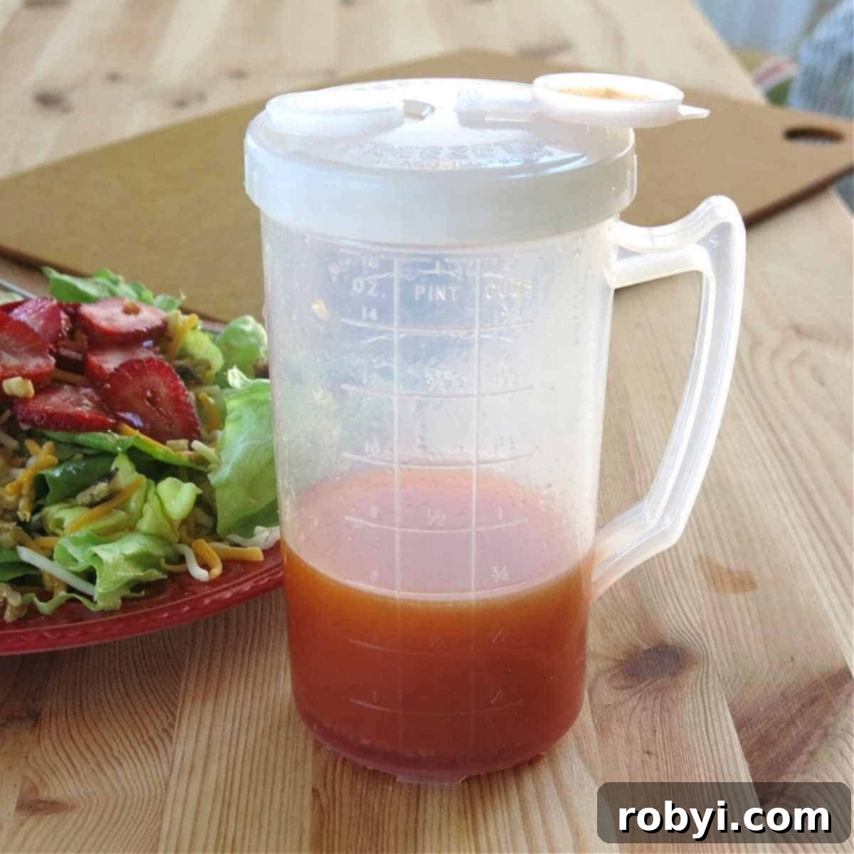 Homemade Red Wine Vinaigrette Dressing in a clear plastic container with measurement markings, sitting alongside a vibrant strawberry salad, highlighting the ease of storage and serving.