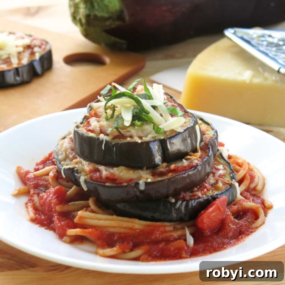 Eggplant Parmesan Stack on a white plate with an eggplant and triangle of Parmesan cheese behind it.