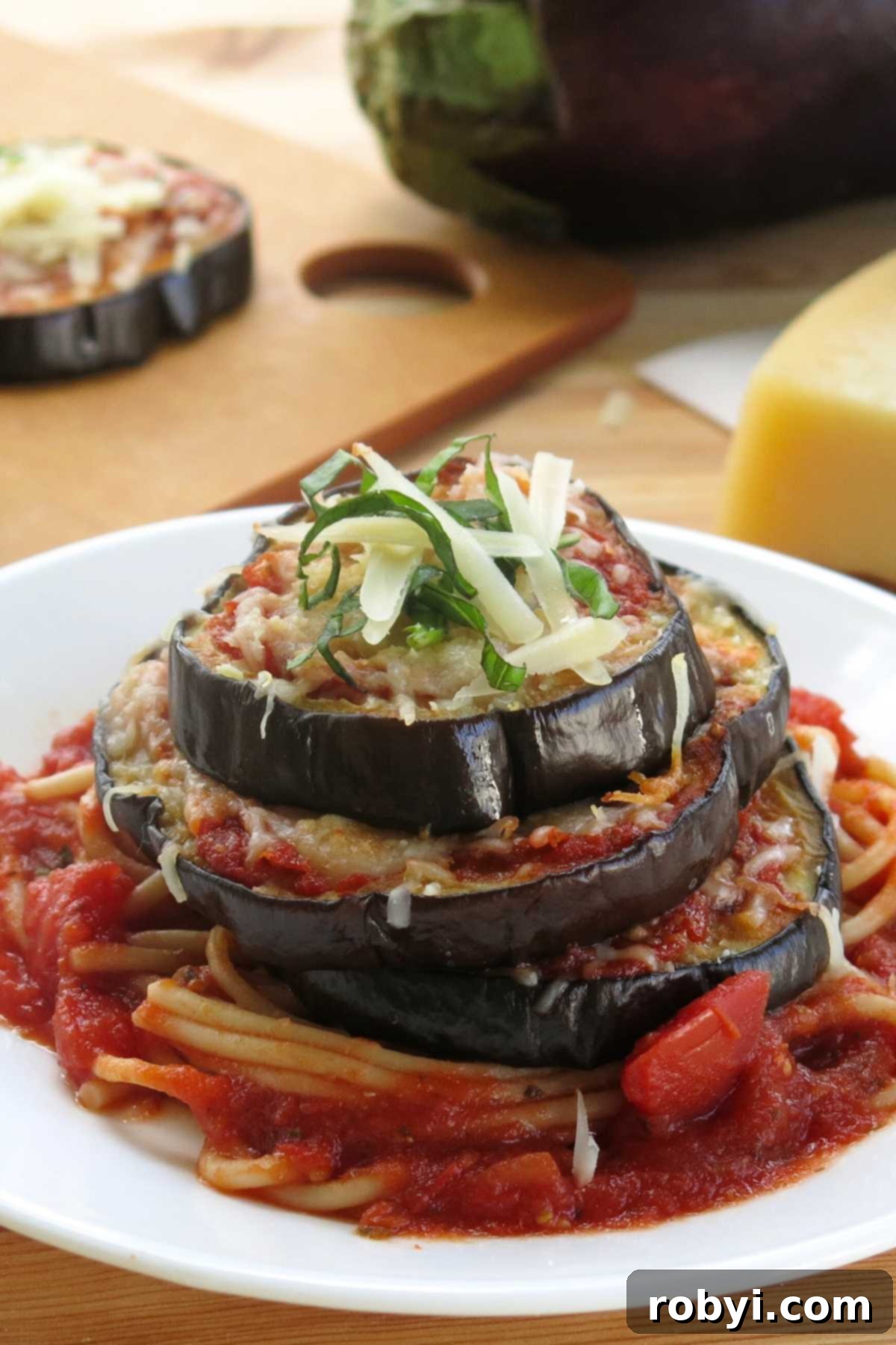 Gluten-free eggplant parmesan stacks on a plate with an eggplant and a cooked eggplant slice behind it.