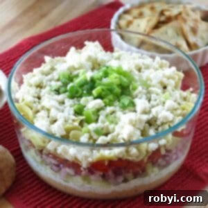 7-layer Greek Dip in a glass bowl with layers of hummus, tsatziki, tomatoes, red onions, feta cheese, and green onions.