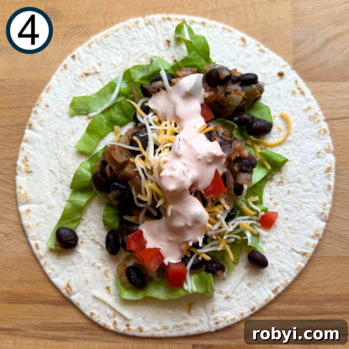 A flour tortilla laid out on a cutting board, with layers of fresh lettuce, black bean mixture, shredded cheese, diced tomatoes, and a dollop of yogurt salsa sauce already added.