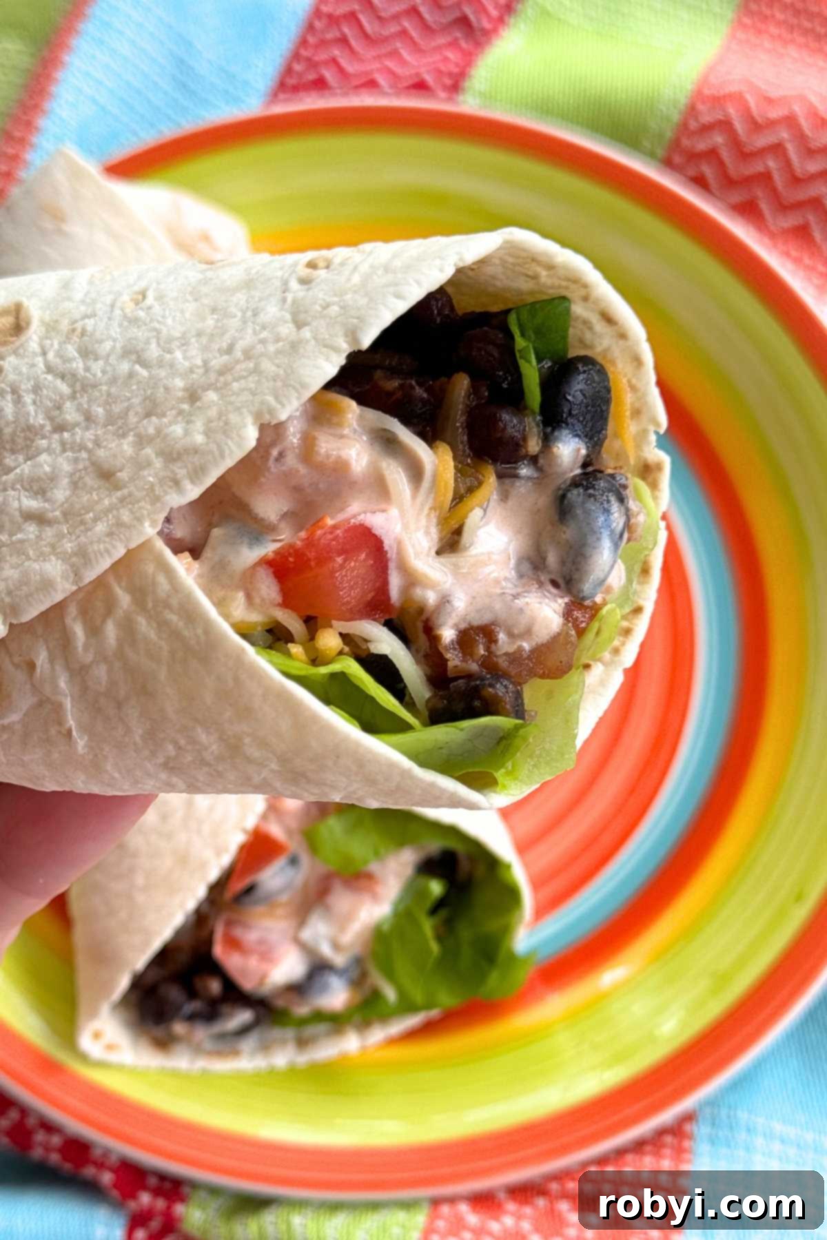 A hand holding a perfectly rolled flour tortilla wrap filled with black beans, cheese, lettuce, tomato, and a drizzle of pink salsa and yogurt sauce. Another identical wrap rests on a colorful plate in the background.