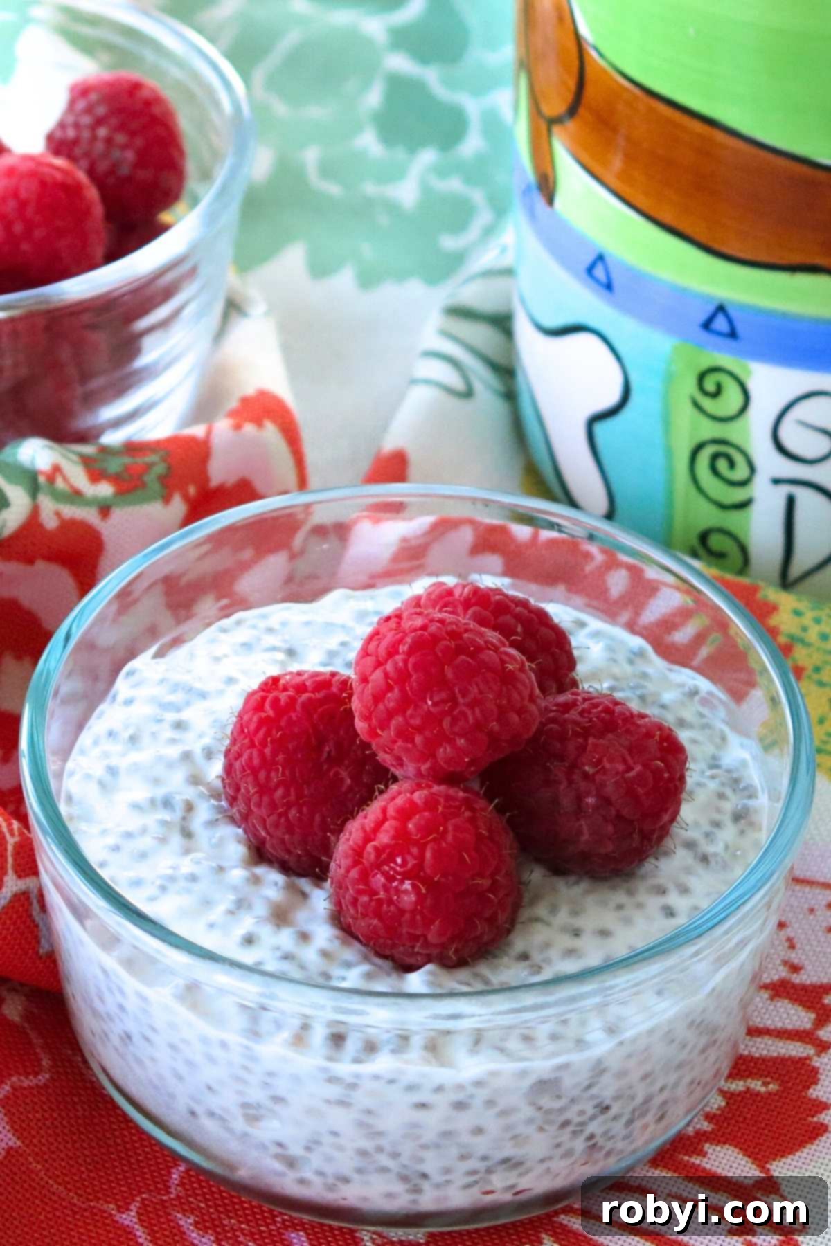 Overnight yogurt chia seed pudding in a glass bowl topped with raspberries with a coffee mug and bowl of raspberries behind it.