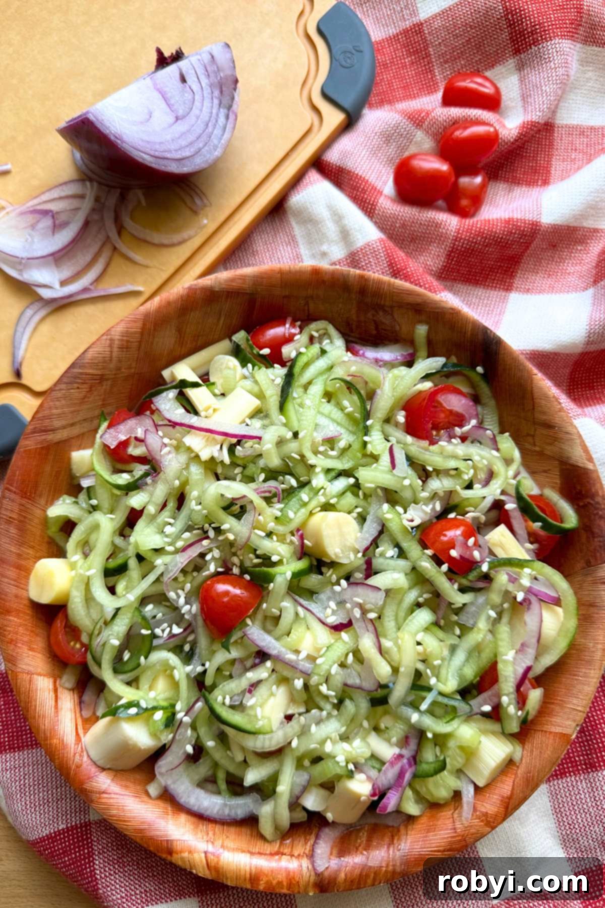 Rice vinegar cucumber salad with red onion, grape tomatoes, hearts of palm, and sesame seeds in a brown bowl with grape tomatoes, and slices of red onion behind it.