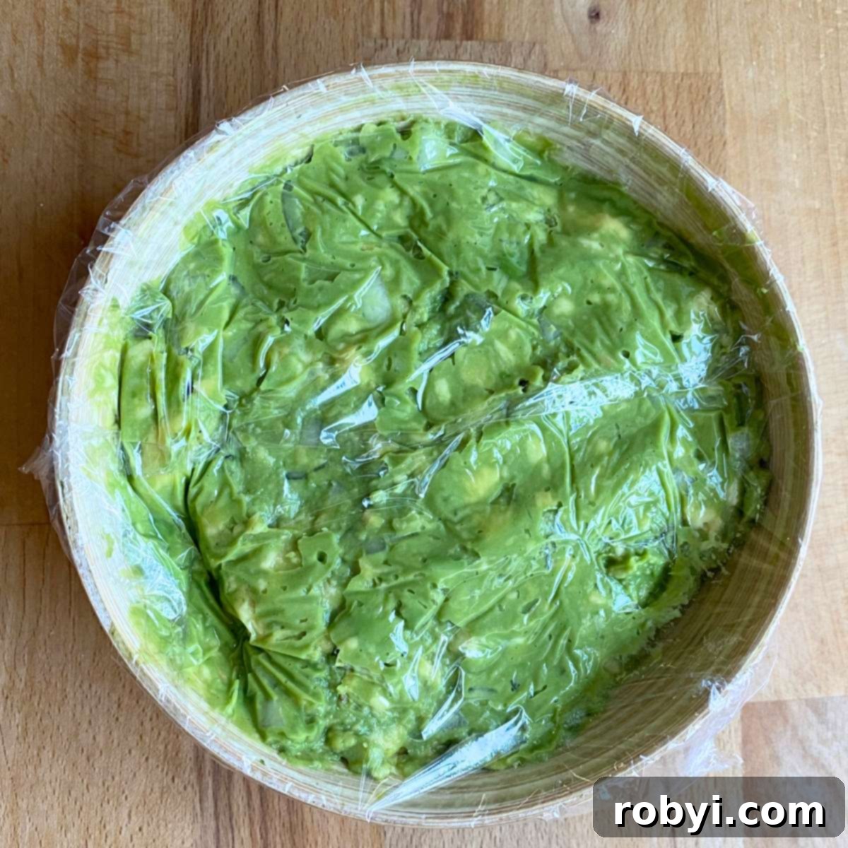 Simple guacamole dip in a bowl with plastic wrap covering the surface for storage.