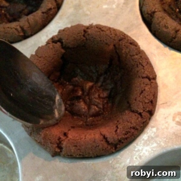 A spoon gently pressing down the sides of a partially cooked cookie cup, shaping it into a deeper, more defined vessel.