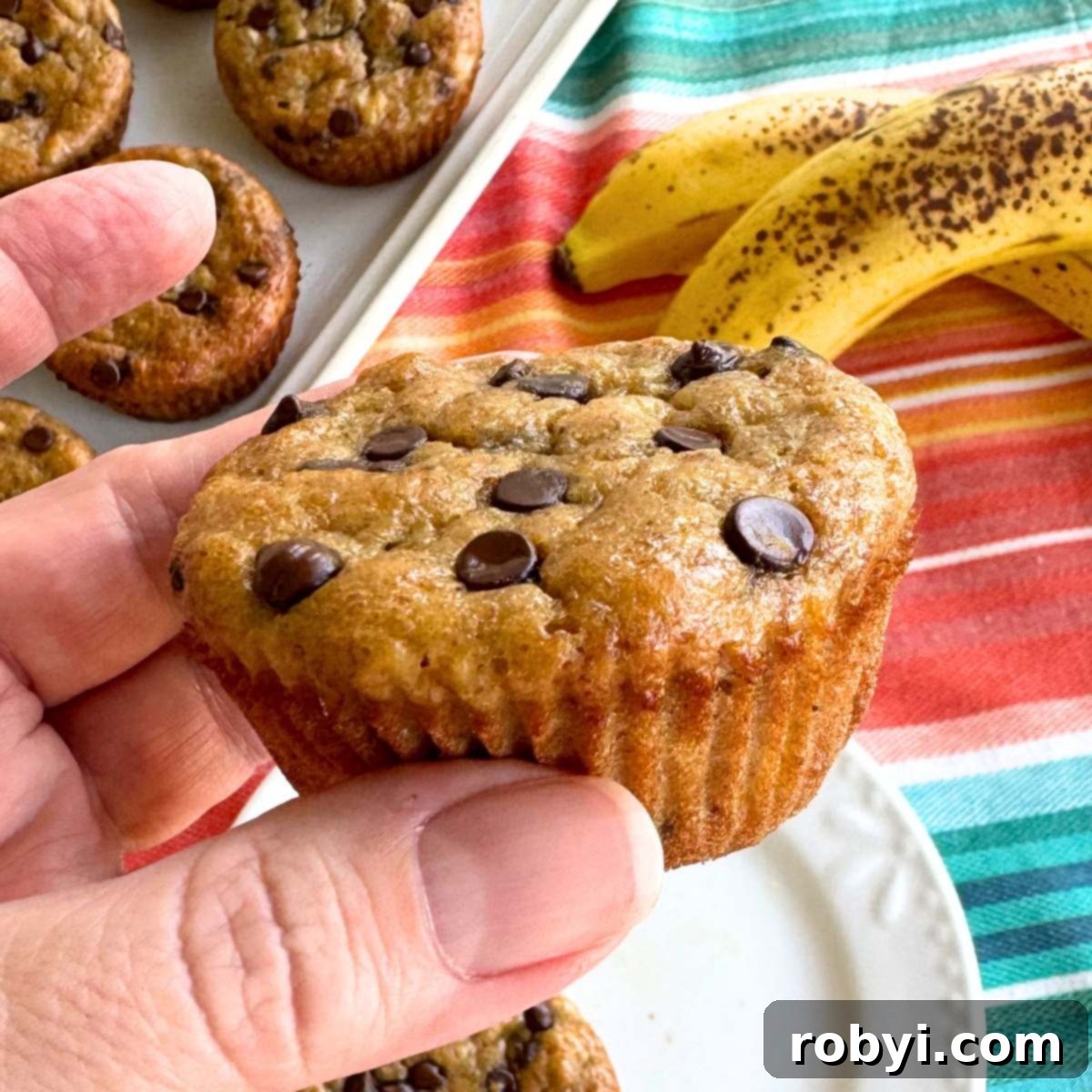 Hand holding a healthy banana yogurt muffin with ripe bananas and more muffins on a platter behind it, emphasizing its delicious and wholesome nature.