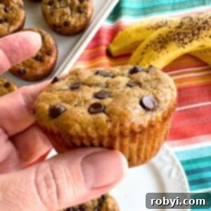 Hand holding a Greek yogurt banana muffin with two ripe bananas and more muffins behind it.