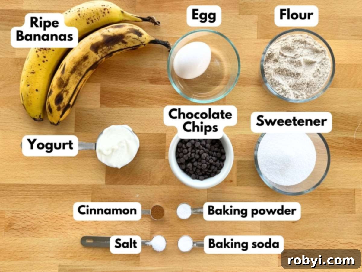 Two ripe bananas, egg, flour, Greek yogurt, mini chocolate chips, sugar and stevia blend, cinnamon, baking powder, baking soda, and salt neatly arranged on a cutting board, ready for baking.
