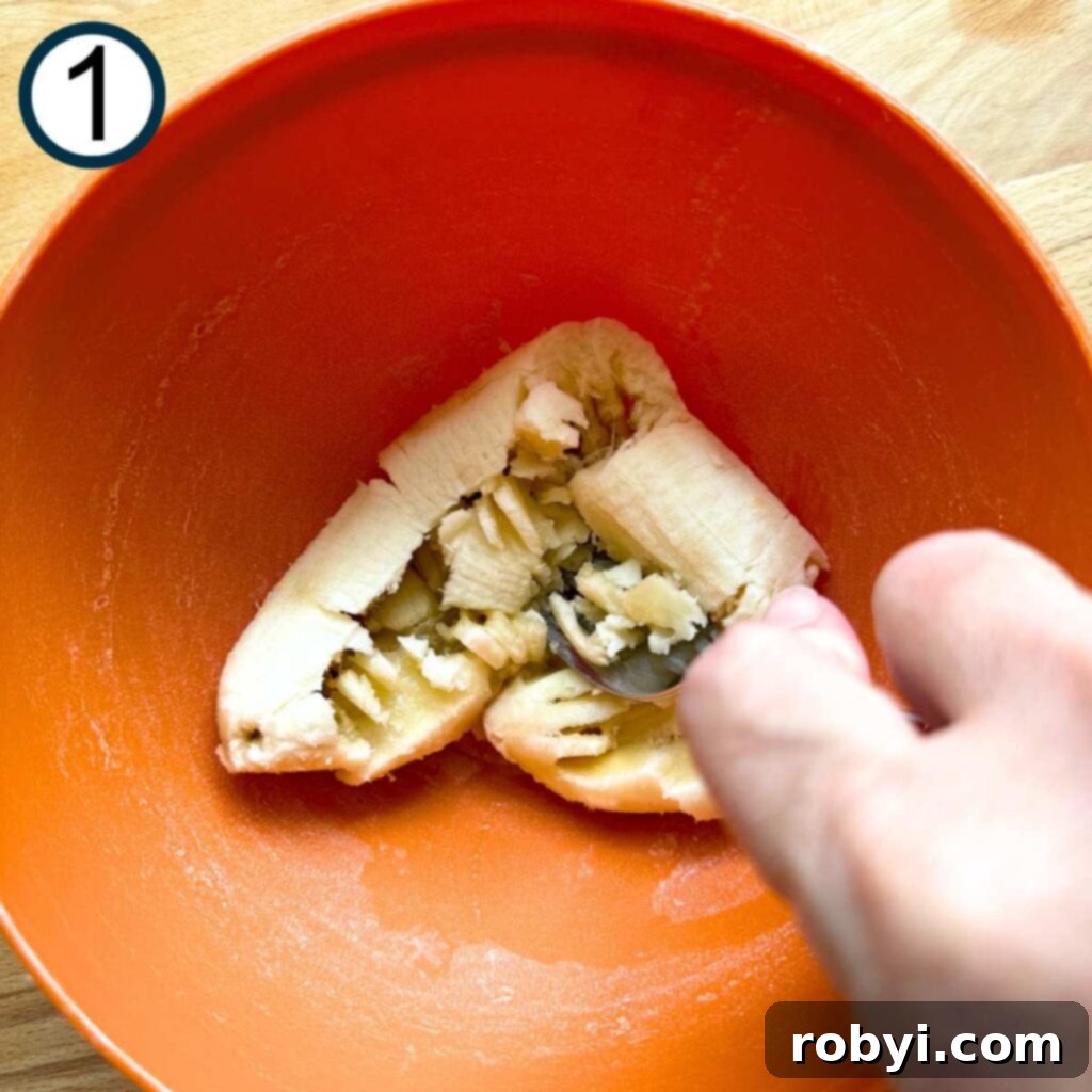 Bananas being mashed with a fork in an orange bowl, showing the texture desired for the muffins.
