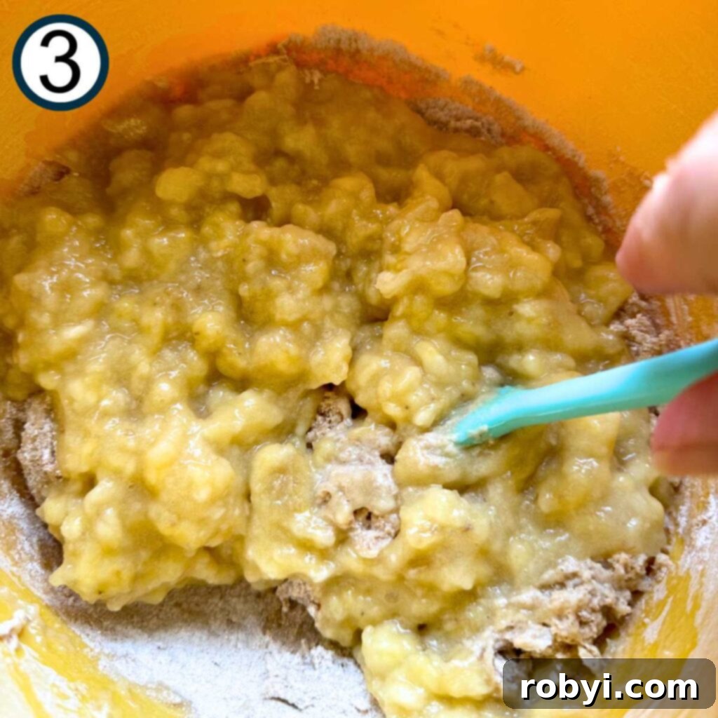 Mashed bananas being stirred into the dry mixture with a spatula, showing the gentle mixing technique.