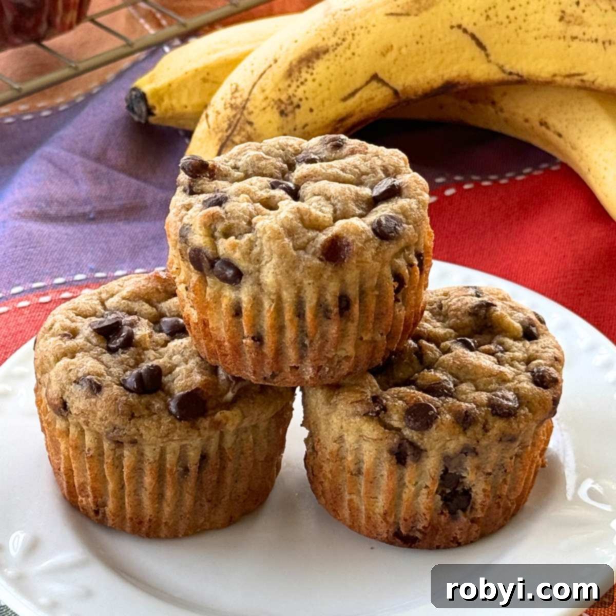 Three Greek yogurt banana muffins stacked in a pyramid on a white plate with two ripe bananas behind it, ready to be enjoyed.