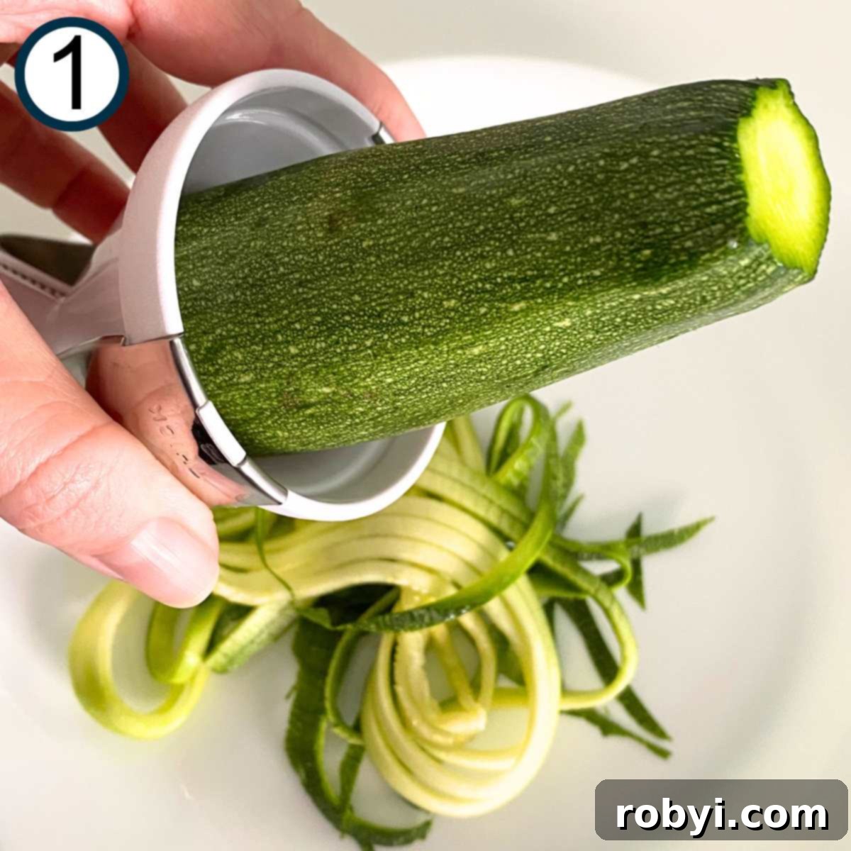 A hand holding a spiralizer, actively transforming a fresh zucchini into delicate spiral noodles, which collect on a white plate below.