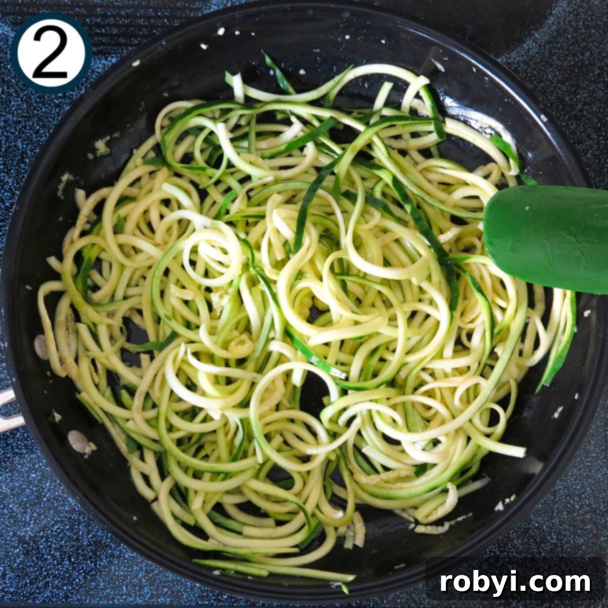 Spiralized zucchini noodles being sautéed in a large skillet, stirred with a green spatula, preparing to soften for the Alfredo sauce.