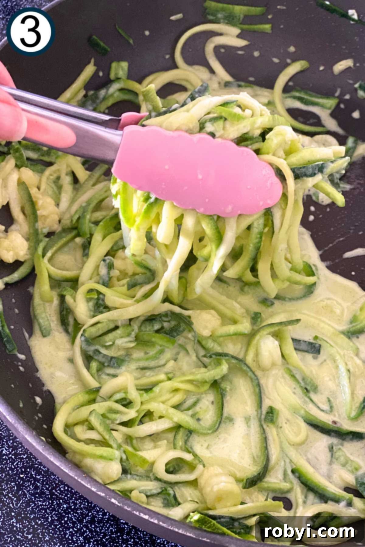 Tongs gently lifting cooked zucchini noodles coated in Alfredo sauce from a skillet, showing the creamy texture and minimal excess sauce.